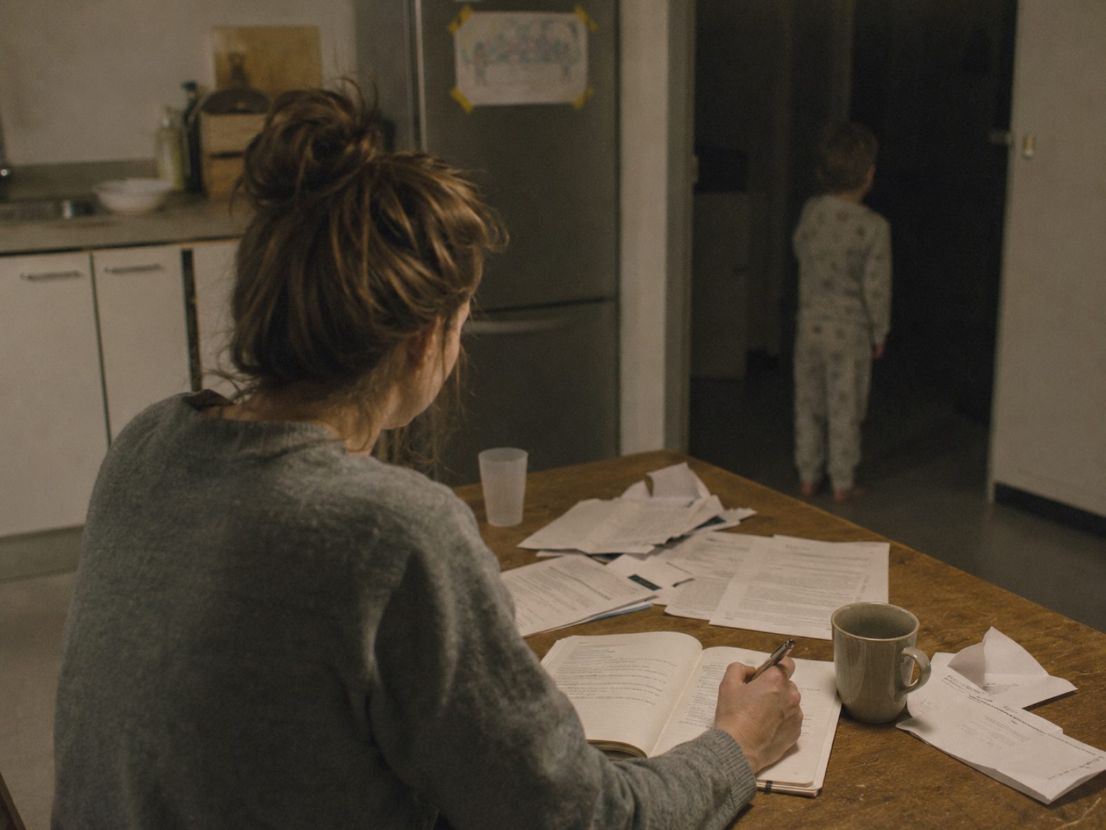 Een moeder zit 's avonds aan de keukentafel tussen brieven en formulieren en schrijft in een notitieboek; achter haar staat haar kind in pyjama in de donkere deuropening