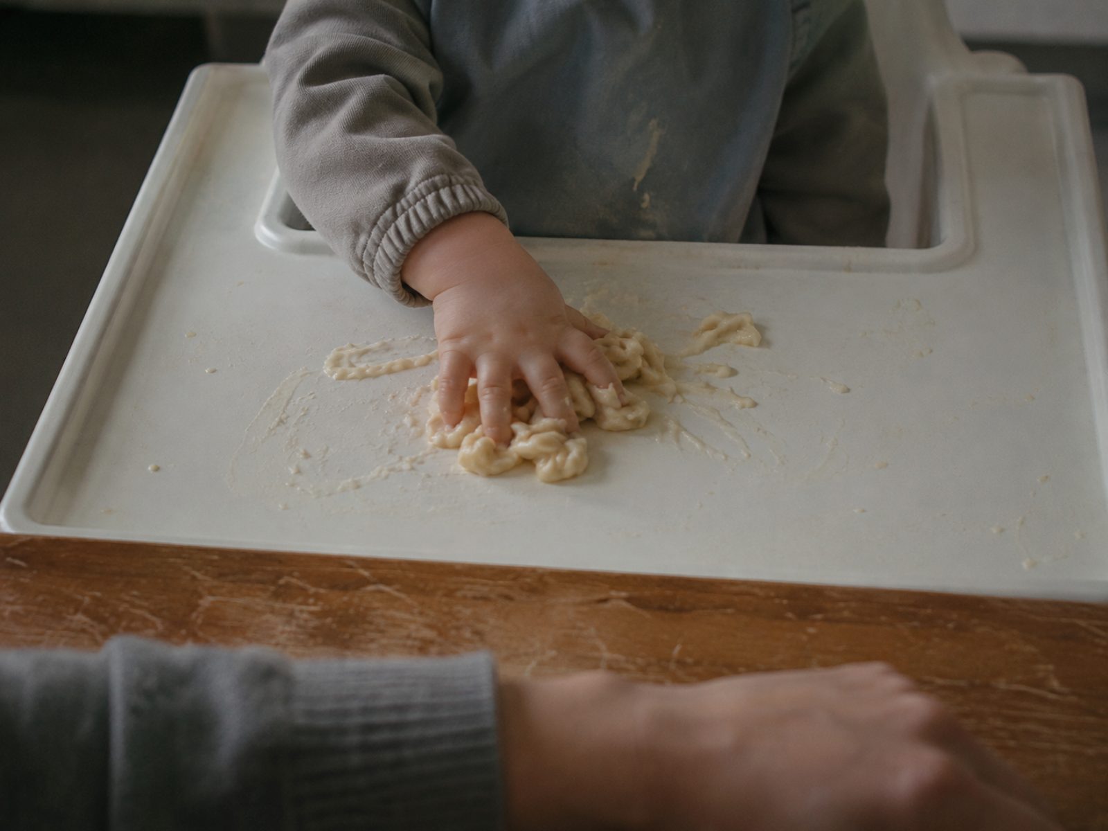 Een klein kind knijpt met de hand in eten op het blad van een kinderstoel; op de tafel ernaast rust de hand van een volwassene, zonder in te grijpen