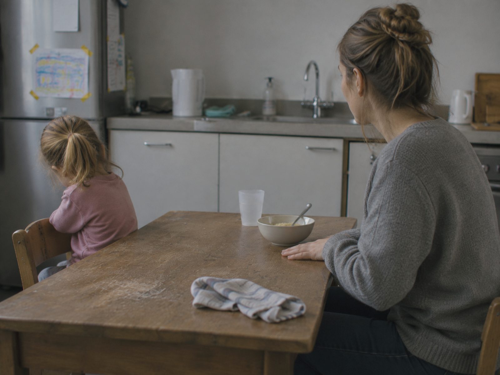 Een meisje heeft zich weggedraaid van de tafel; aan de overkant staat een schaaltje bij de hand van haar moeder, die niets aanreikt